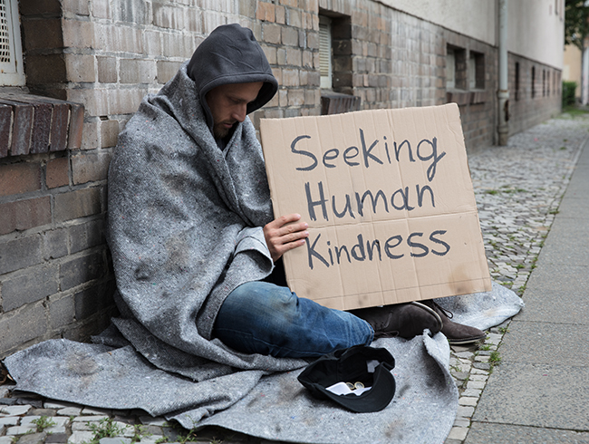 Male Beggar In Hood Showing Seeking Human Kindness Sign On Cardboard