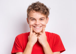 Portrait of teen boy showing his smile. Child pointing with fingers teeth and mouth. Caucasian young teenager on grey background.